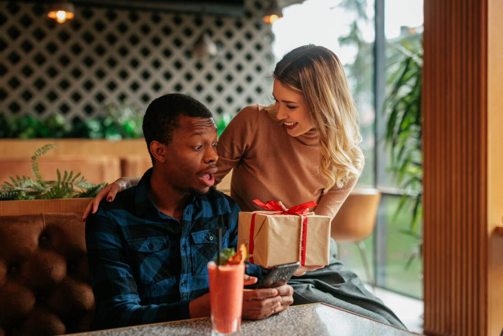 A woman surprises a man with a gift at a cafe.