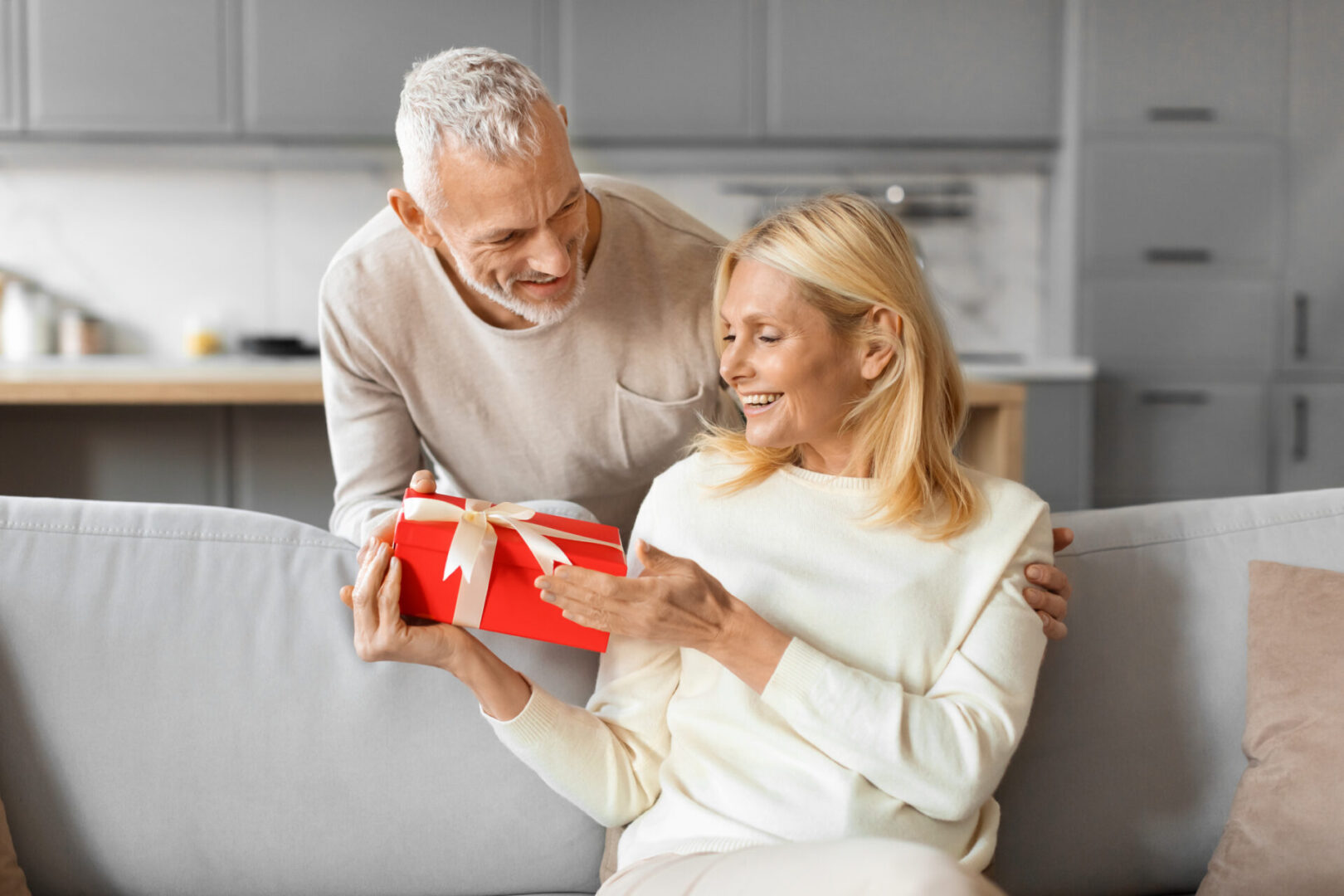 Elderly man giving a wrapped gift to a smiling elderly woman at home.