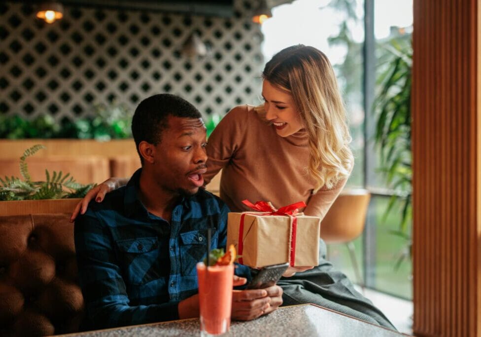 A woman surprises a man with a gift at a cafe.