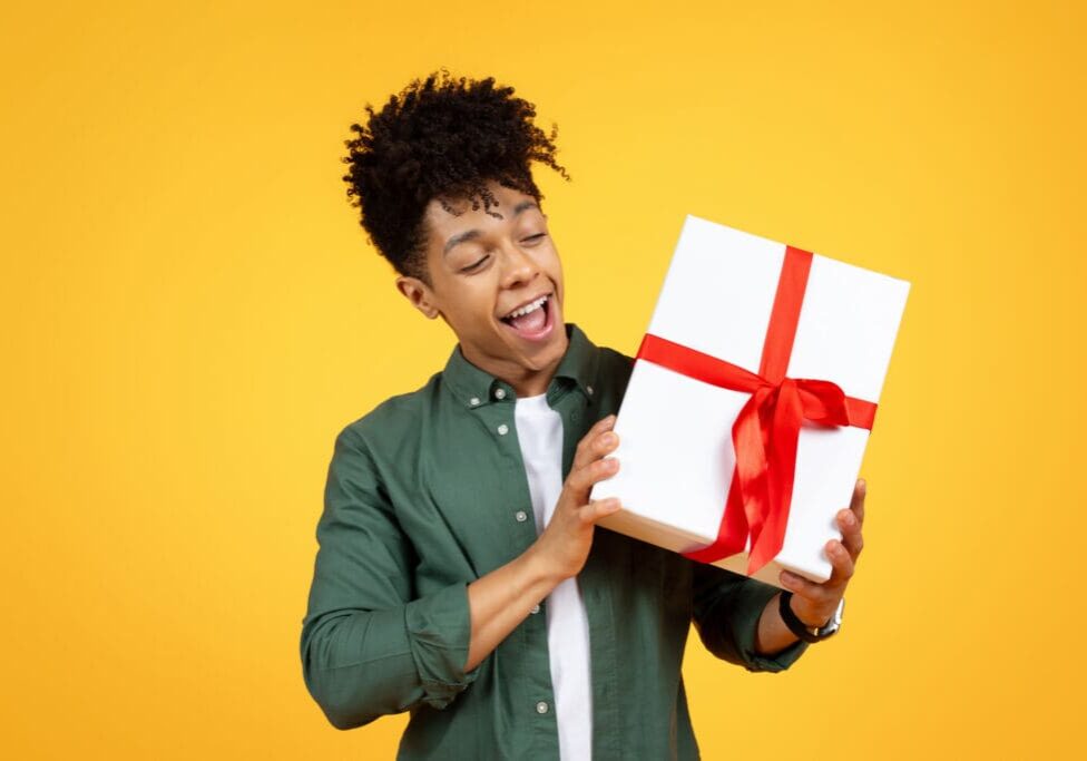 Excited young man holding a gift box with a red ribbon against a yellow background.