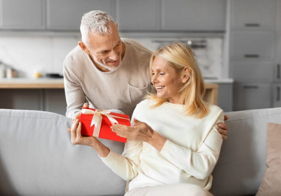 Elderly man giving a wrapped gift to a smiling elderly woman at home.